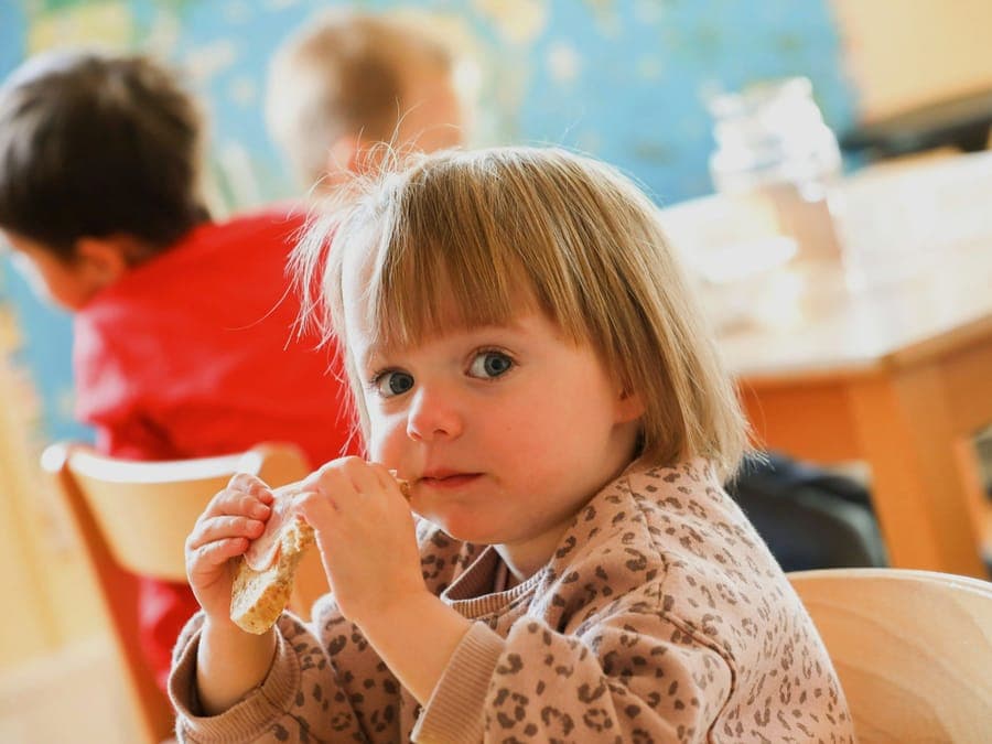 FitKids Kinderkrippe Wiesbaden: Kleinkind mit einem Brot in der Hand schaut in die Kamera