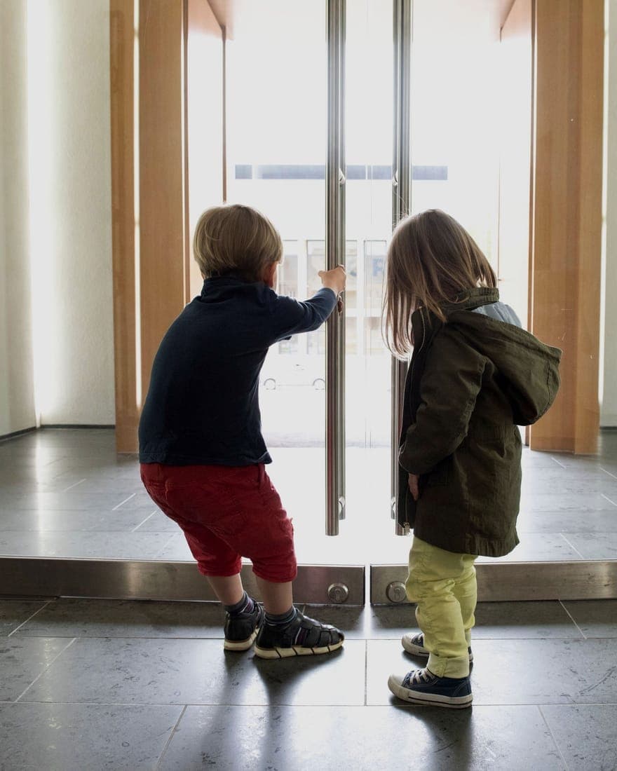 Zwei Kinder schauen von innen durch die große Glastür einer Einrichtung von Fit for Family Care