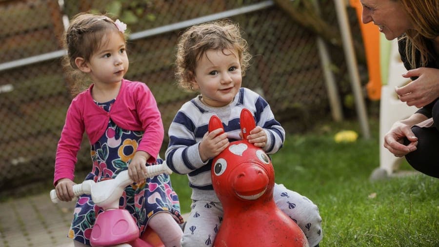 FitKids Kinderkrippe: zwei Kleinkinder auf Spielfahrzeugen im Garten, eine Erzieherin lacht mit ihnen