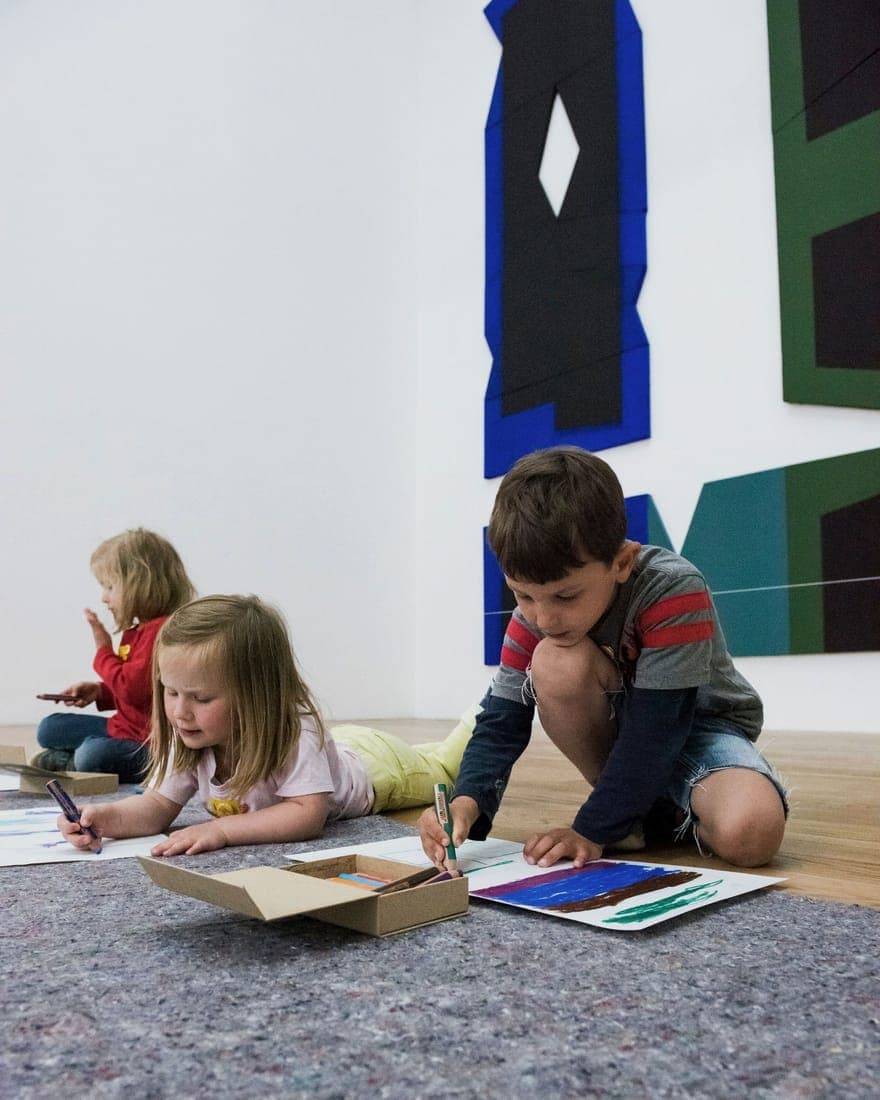 Drei Kinder malen gemeinsam am Boden vor Gemälden im Kunstprojekt