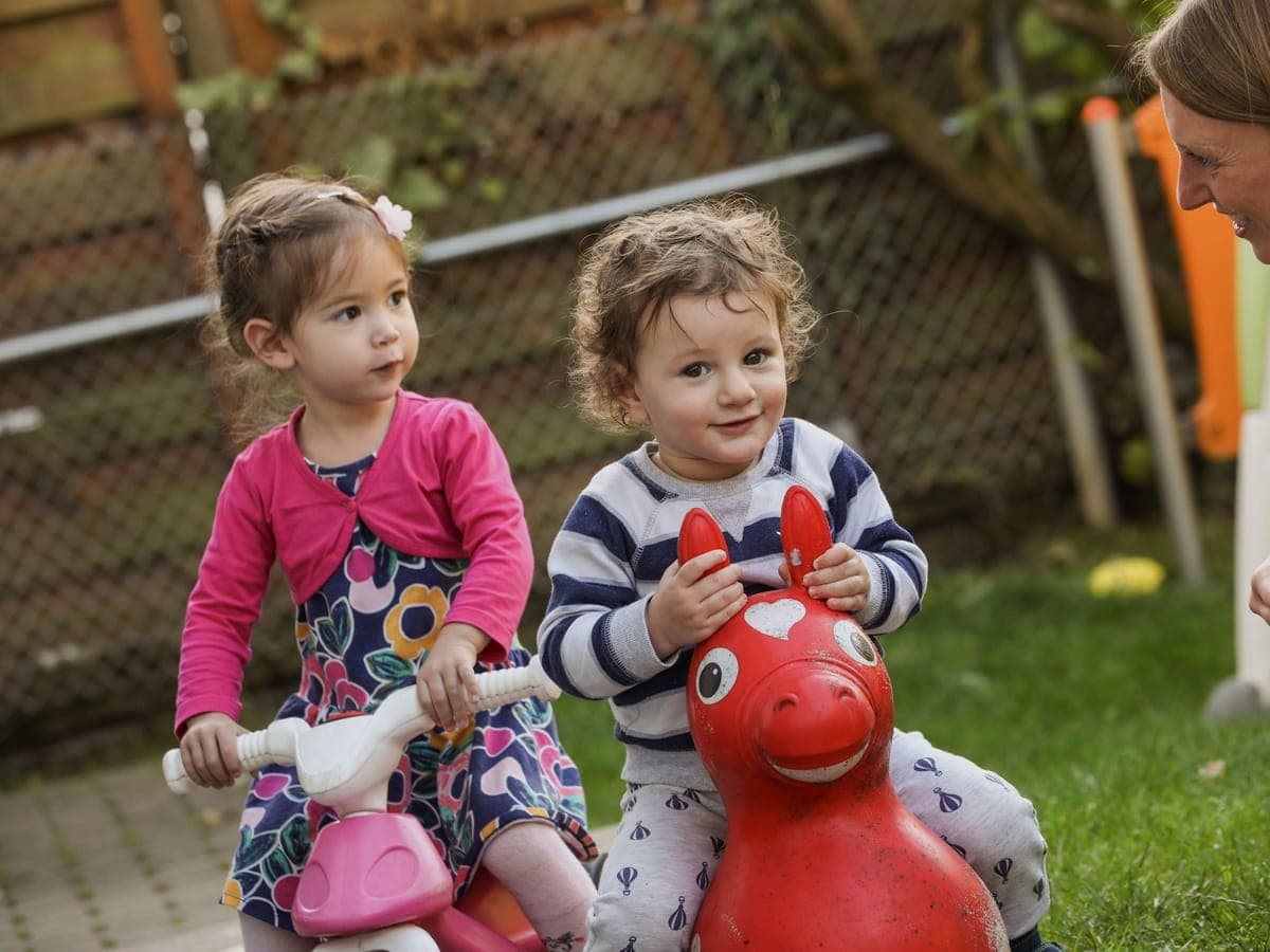 Zwei Kleinkinder auf Spielfahrzeugen, eine Erzieherin lacht mit ihnen